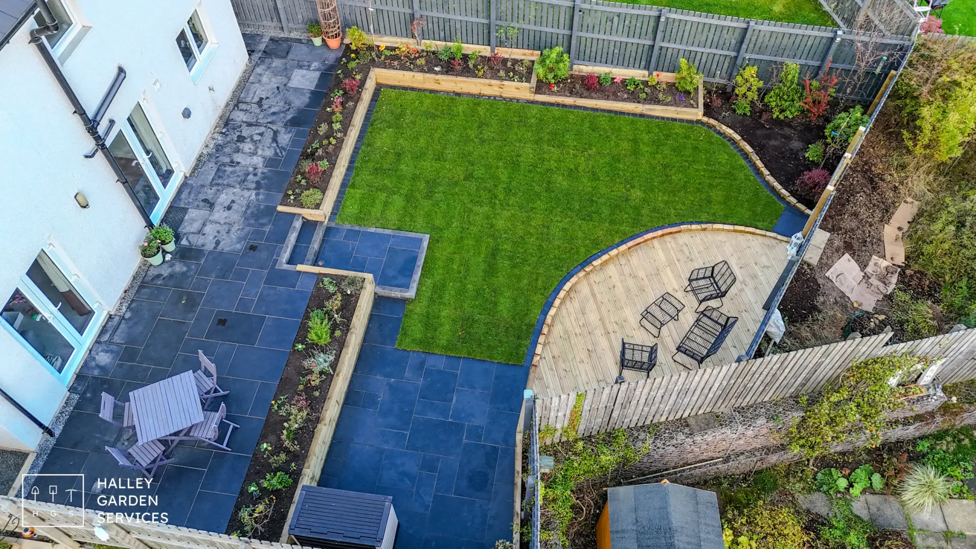 Aerial view of a landscaped back garden featuring a modern garden design with fresh turf, black slate patio with seating, raised flower beds, and a wooden deck area with chairs for outdoor living.