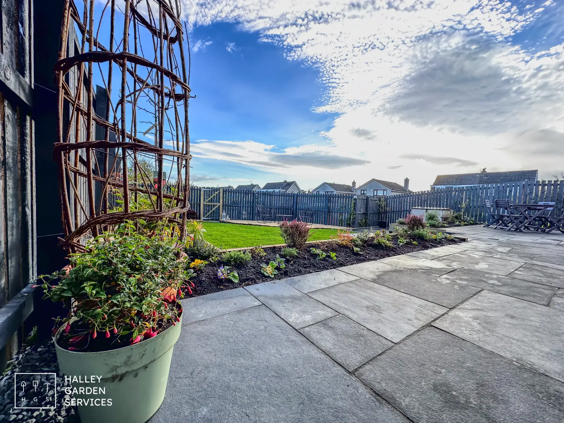 Neatly landscaped back garden with stone patio, potted plant, flower beds, modern garden design elements, green lawn, and wooden fence under a partly cloudy sky in Midlothian