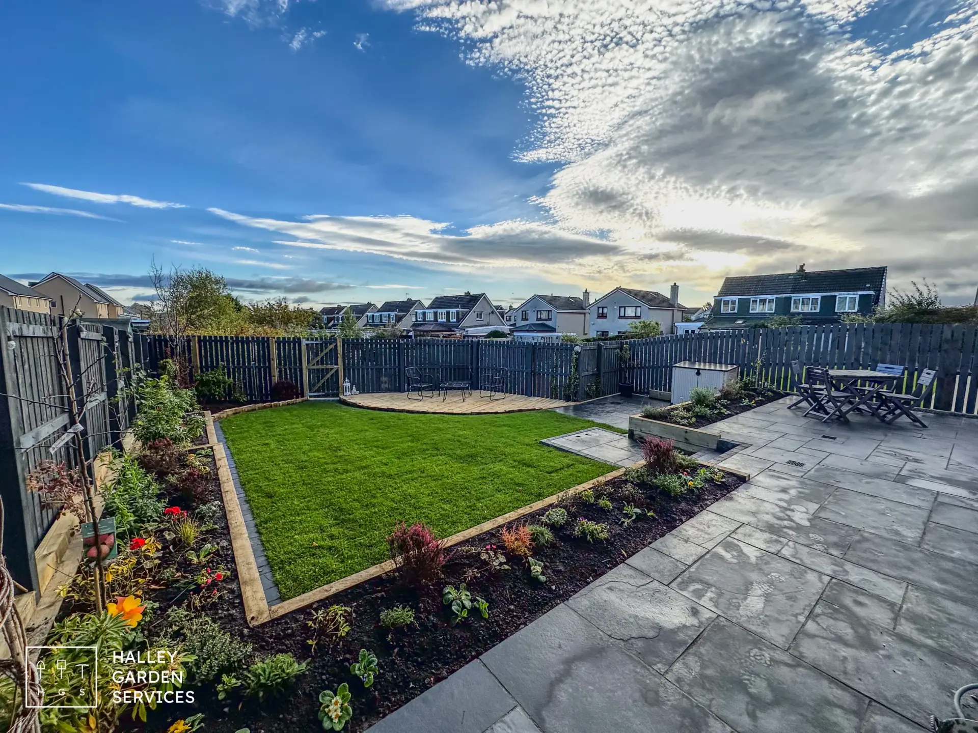 A well-maintained garden bordered by vibrant flowers. Wooden fence encloses the area, with a patio and outdoor furniture in Mauricewood